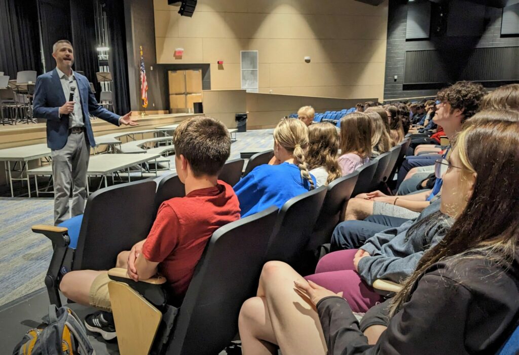 Jake Bleacher speaks in front of an audience of about 100 students in the auditorium.