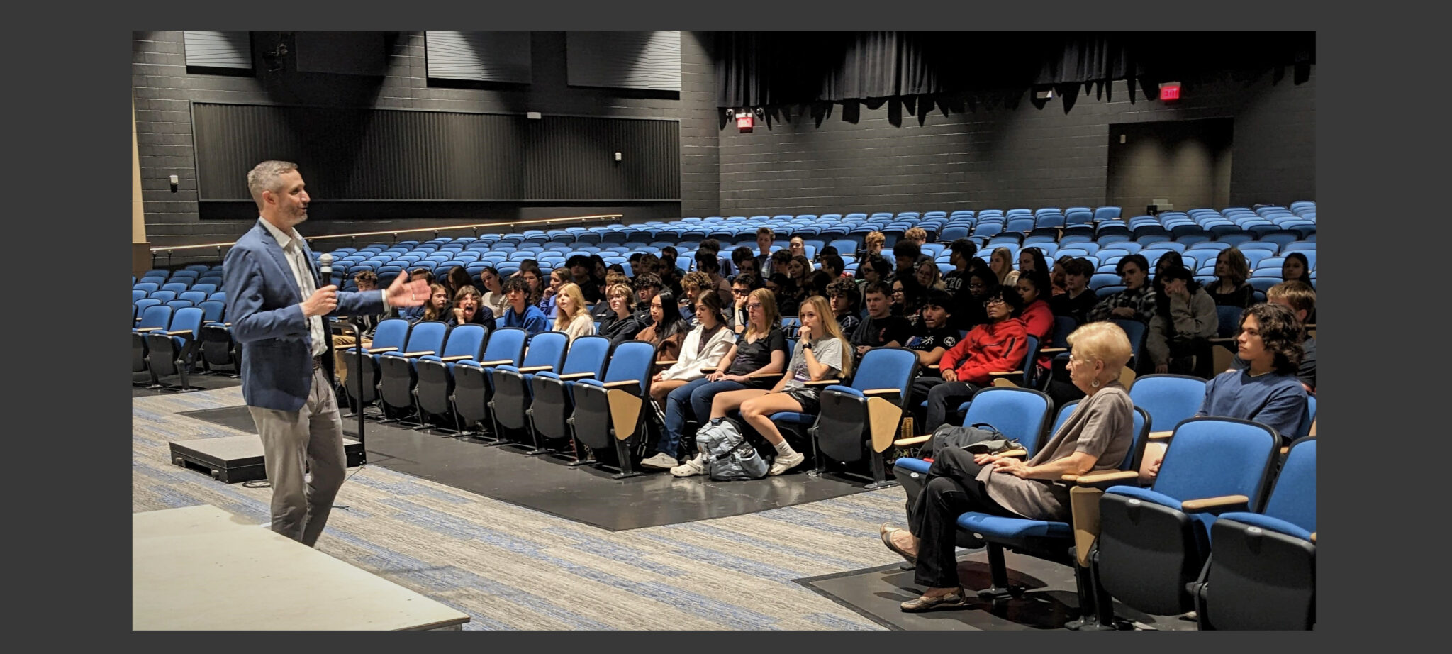 Jake Bleacher speaks in front of an audience of about 100 students in the auditorium.