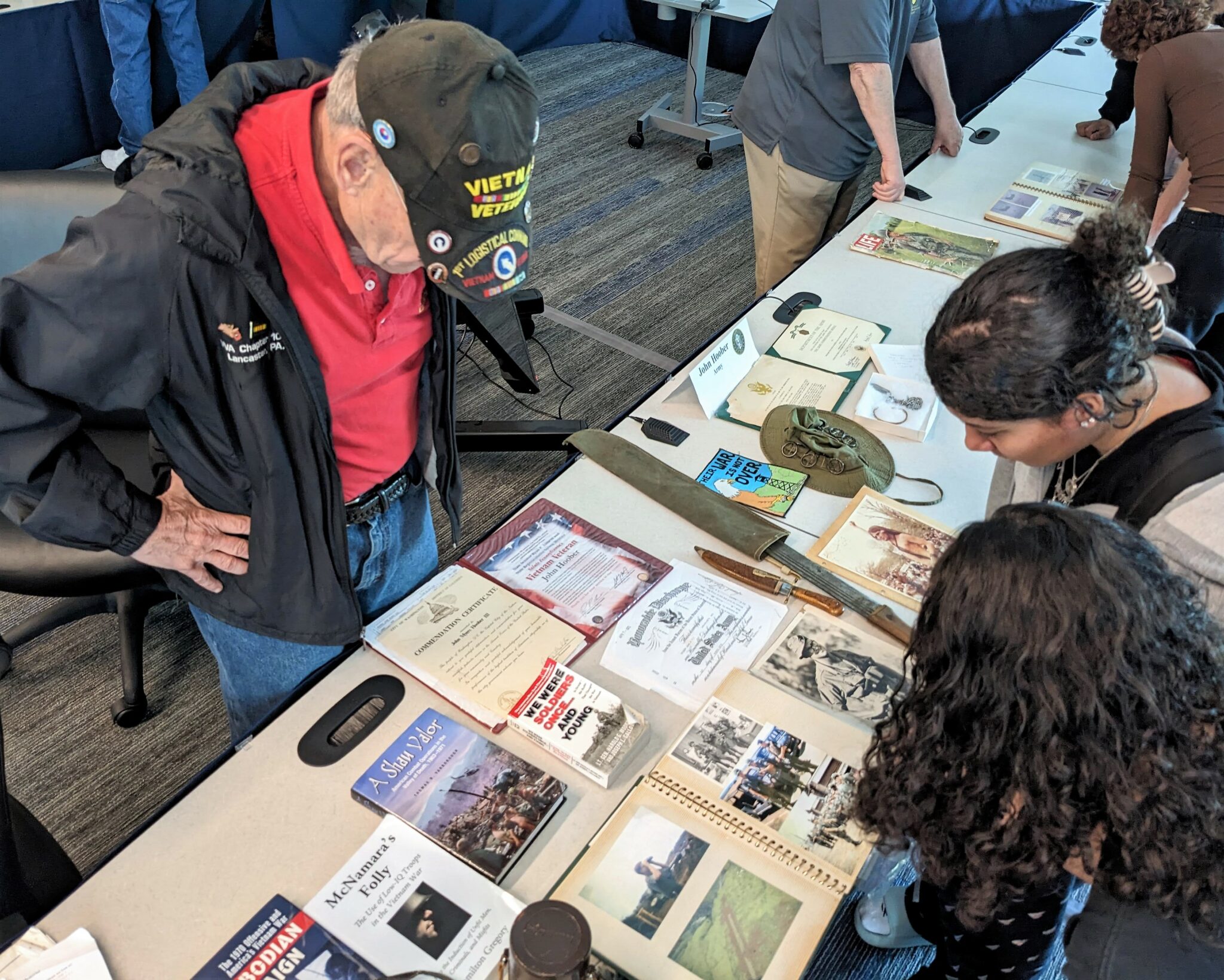 A Vietnam Veteran stands near two students looking at his memorabilia.