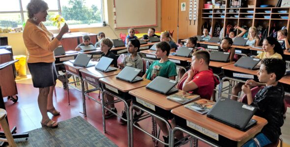 A staff member instructs elementary students using laptops