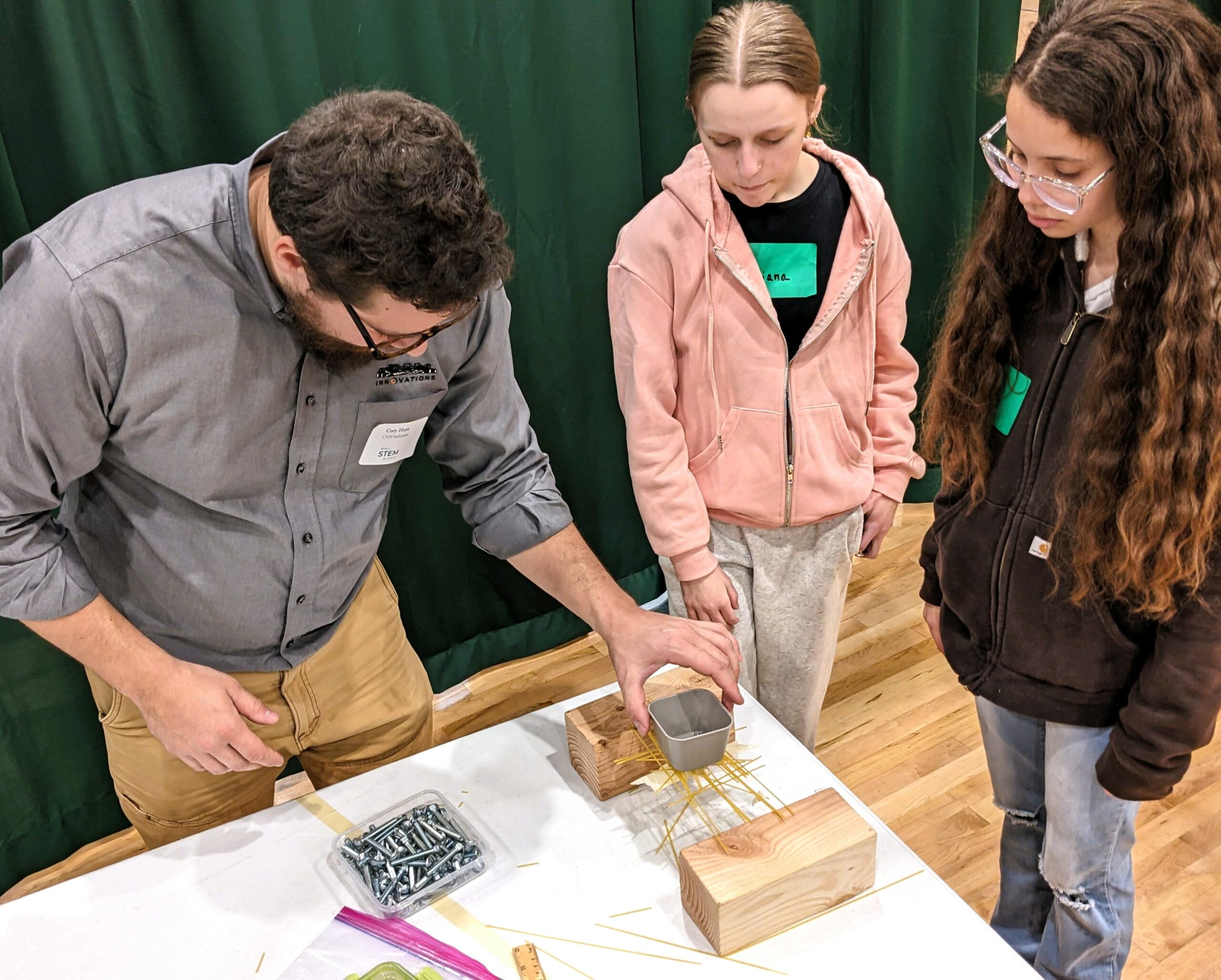 Two students watch as a volunteer tests their spaghetti bridge design.