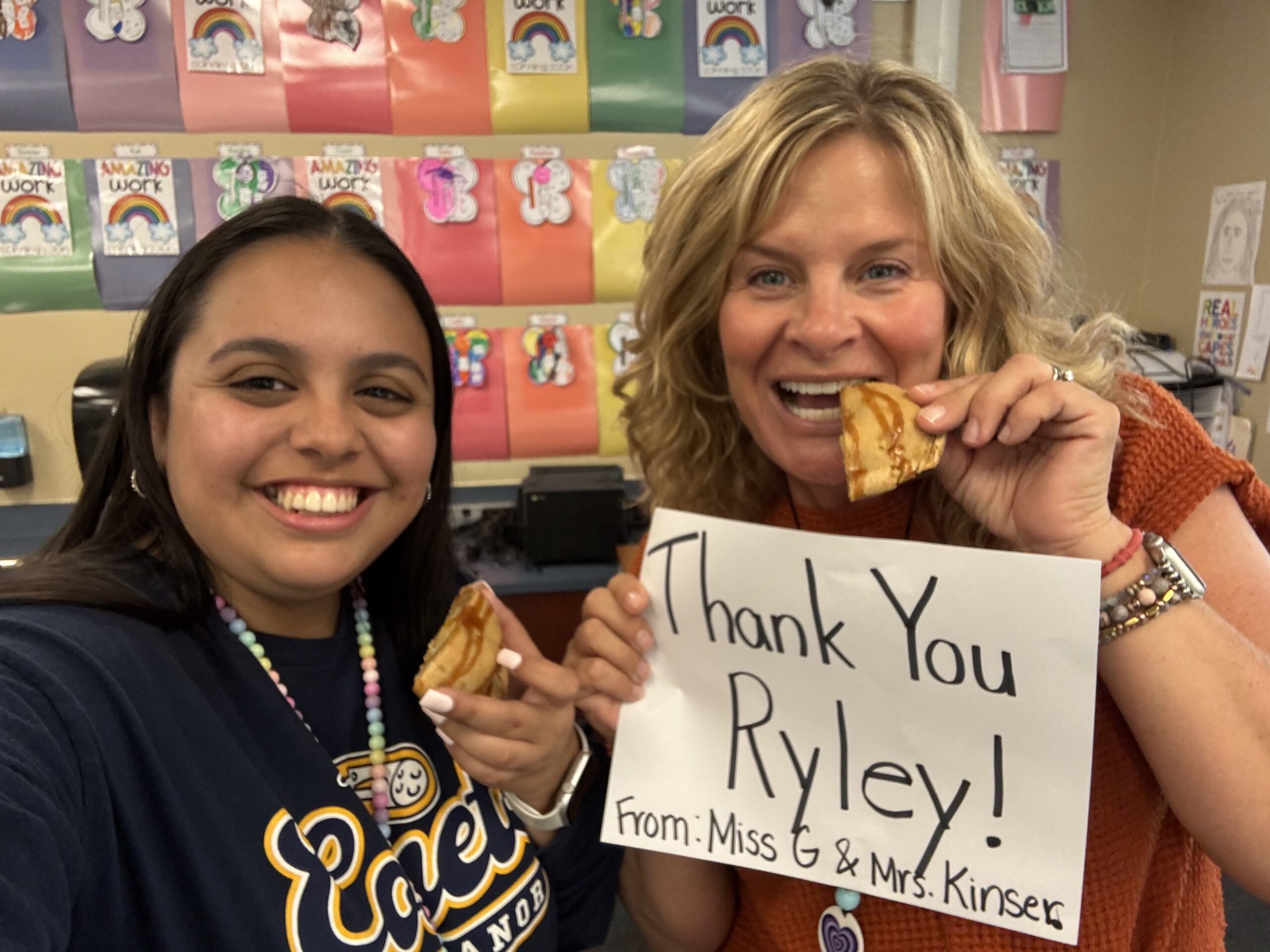 Two teachers eating cookies and holding a "thank you Ryley" sign
