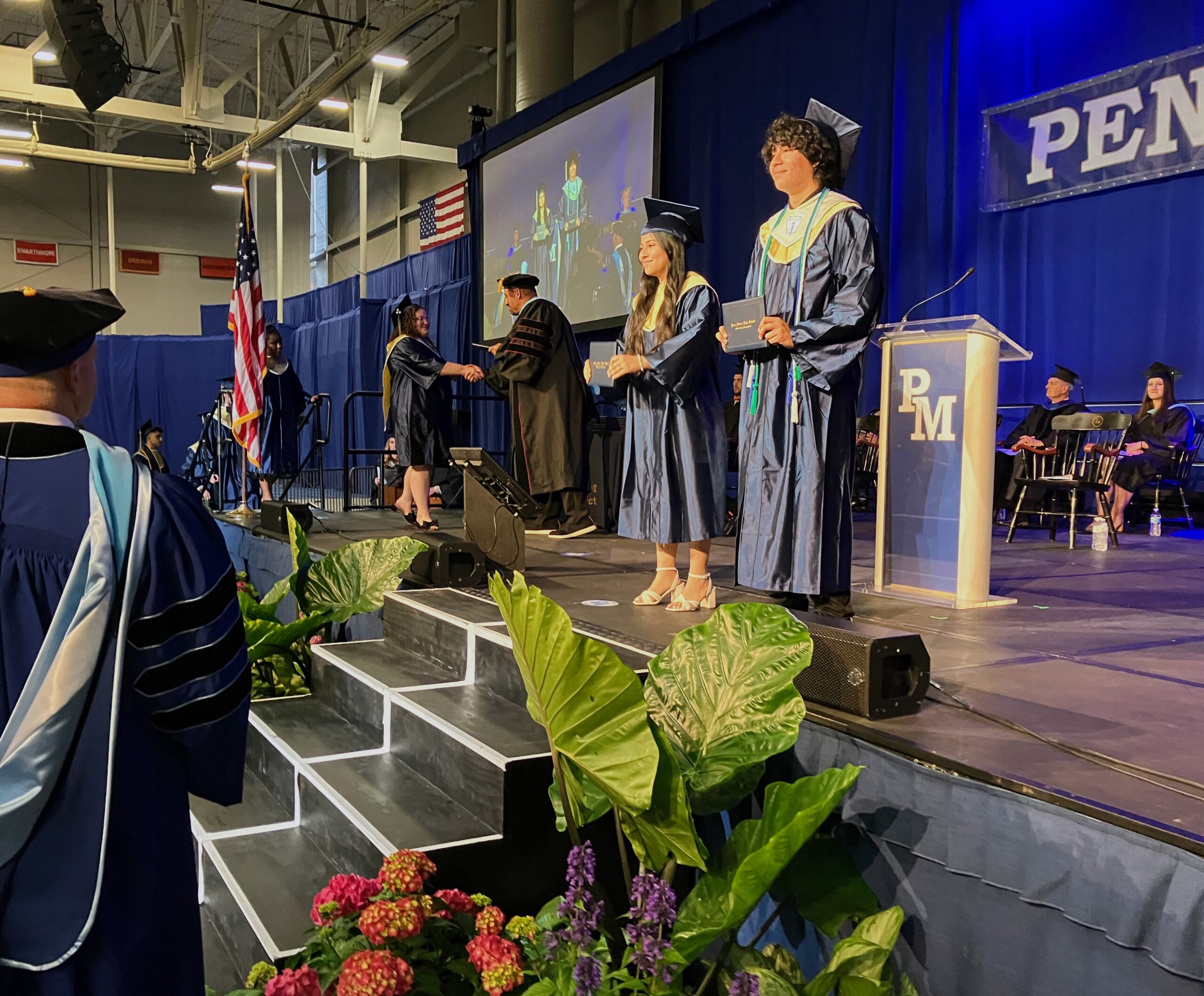 Two graduates stop onstage to pose for a photo with their diplomas.